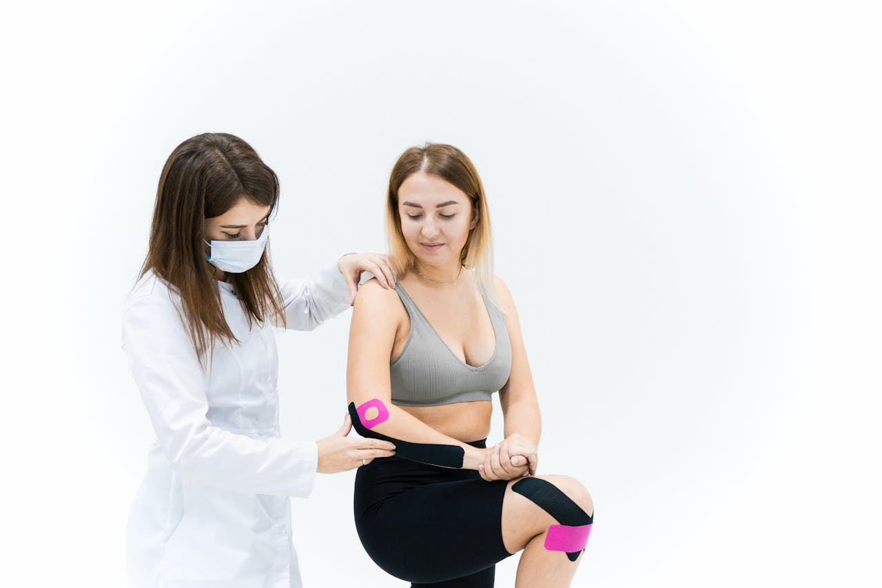 A therapist applies kinesio tape on a woman's knee during a physical therapy session on a white background.